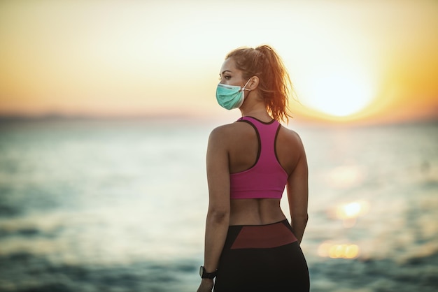 Снимок бегуньи в защитной маске во время тренировки возле пляжа на закате во время пандемии COVID-19.