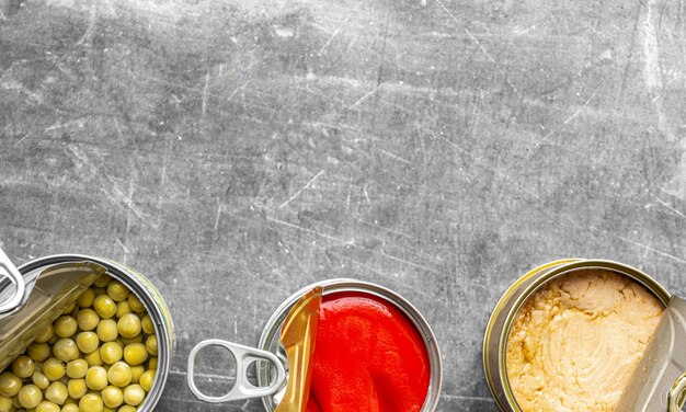 Shot from above of three cans of canning lined up at the bottom of the open image of red pepper, light tuna and peas with gray surface and soft natural lighting