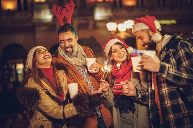 저녁 파티에서 크리스마스 시장에서 폭죽과 함께 즐거운 시간을 보내고 뜨거운 음료를 즐기는 쾌활한 젊은 친구들의 사진.