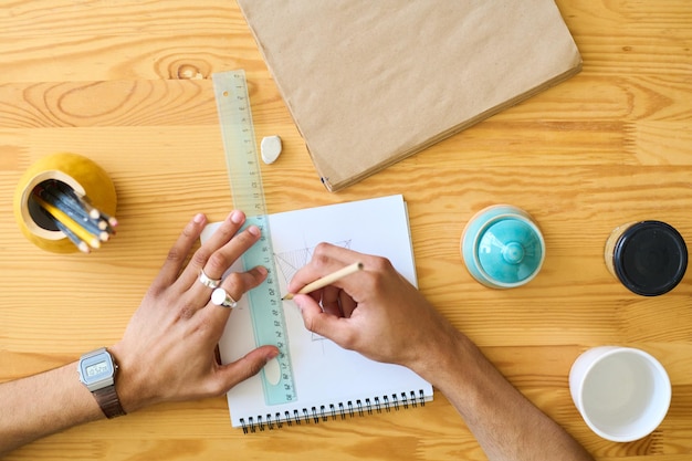 Above shot of hands of multiethnic guy making sketch of new item of earthenware