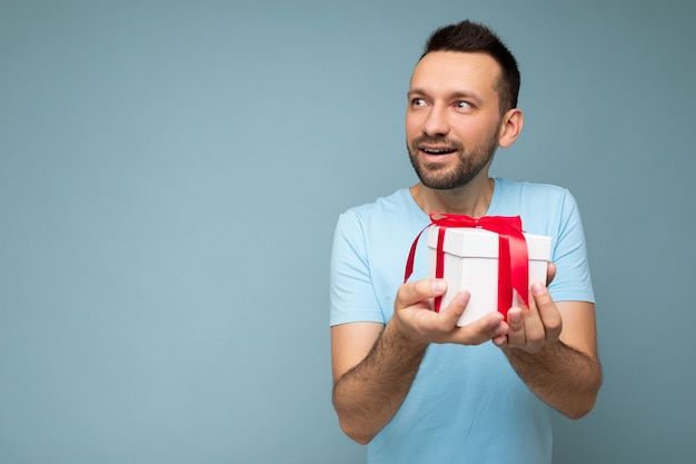 Shot of handsome happy smiling brunet young male person with beard isolated over blue background wall wearing blue t-shirt holding white gift box with red ribbon and looking to the side. Copy space