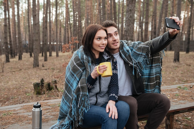 shot of handsome man with his smiling woman with cup making selfie using smartphone in wood