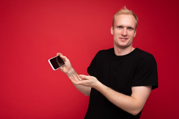 Photo shot of handsome smiling blonde young male person wearing black t-shirt standing isolated on red background with copy space holding smartphone showing phone in hand with empty display for mockup point