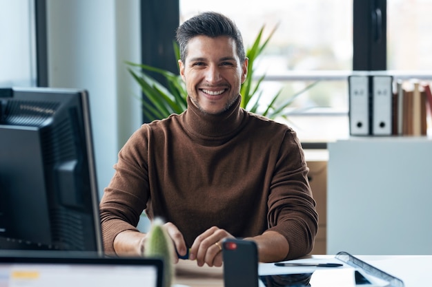 Shot of handsome young entrepreneur looking at camera while using the computer in modern startup office.