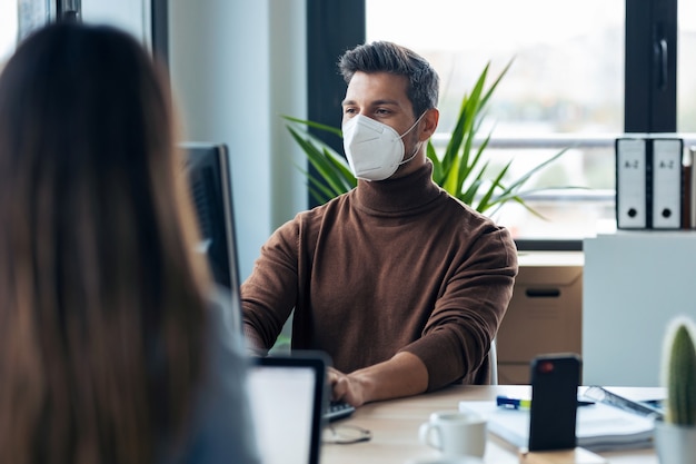 Shot of handsome young entrepreneur wearing a hygienic face mask working with computer in modern startup office.