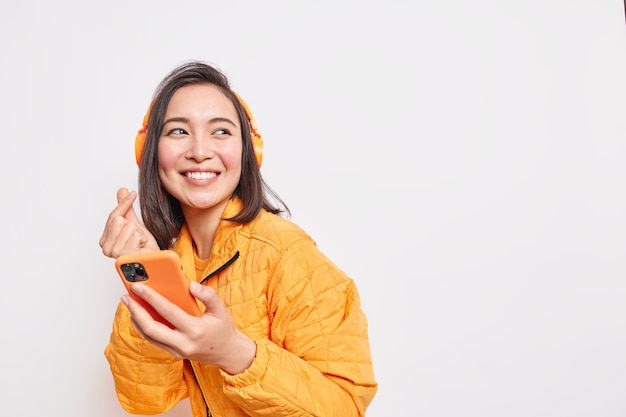 shot of happy Asian millennial girl makes eastern like sign smiles gladfully looks away dressed in orange jacket uses smartphone and headphones for listening favorite music poses indoor