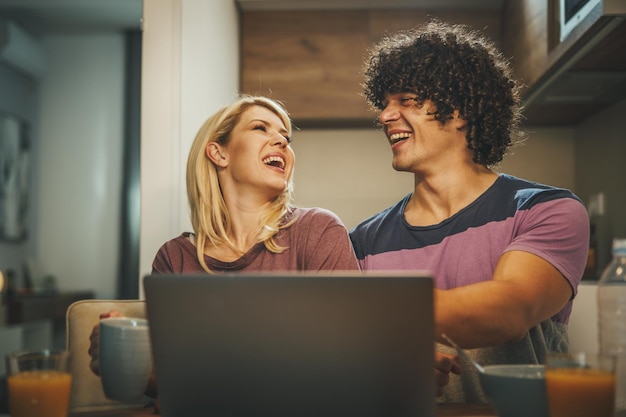 Photo shot of a happy couple using laptop together and eating breakfast before work in the kitchen.