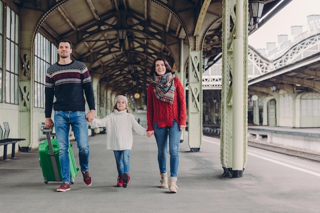 Shot of happy family going to have nice trip during holidays carry bag walk on railways station platform being in good mood Father mother and child arrive from journey Travelling concept