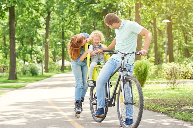 Colpo di genitori felici coccole con il loro bambino mentre in bicicletta e pattinaggio insieme al parco amore affetto famiglia affetto genitori emozioni infanzia stile di vita attivo.