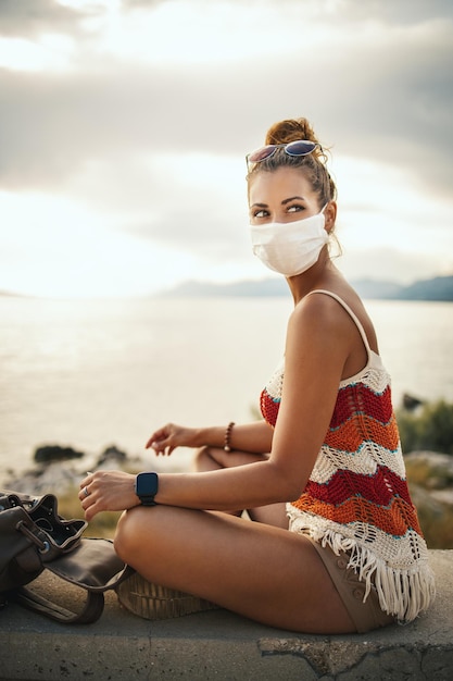 Shot of a happy young woman with protective mask spending time\
on seaside during exploring a mediterranean at corona\
pandemic.