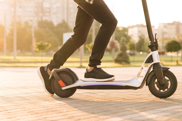 Photo shot on legs young man in black sneakers ride on electric mobile scooter. close up feet man courier food delivery rides the street on electric scooter deliver online order client at sunset