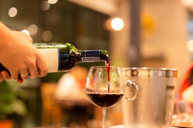 Girato in mano con poca luce versando vino rosso dalla bottiglia in un bicchiere da vino.