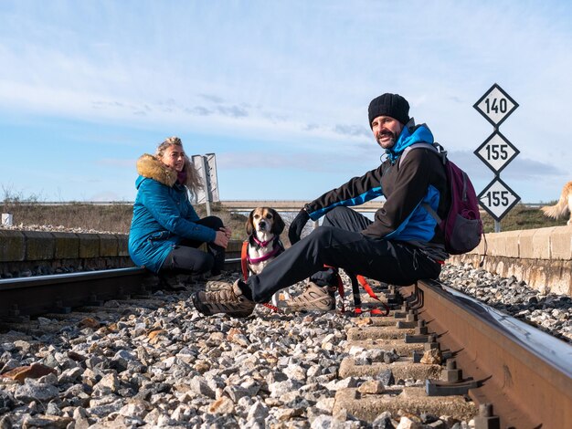 Снимок мужчины и женщины, сидящих на железной дороге со своей очаровательной собакой под ясным небом.