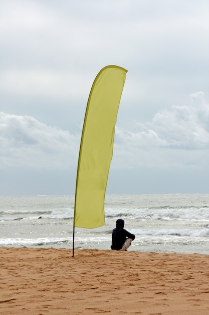 大きな旗の横にある波を見ているビーチの上に座っている男のショット。