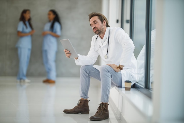 Covid-19パンデミック時に病院の廊下の窓の近くに座っているときにデジタルタブレットを使用している成熟した医師のショット。