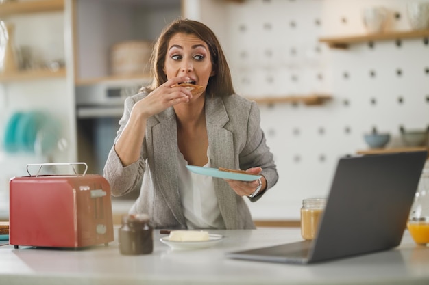 Inquadratura di una giovane donna d'affari multi-tasking che fa colazione e usa il laptop nella sua cucina mentre si prepara per andare al lavoro.