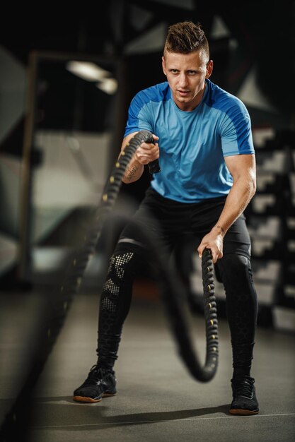 Снимок мускулистого парня в спортивной одежде, делающего упражнения с веревками в спортзале.