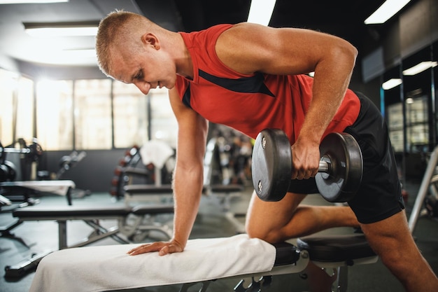 Снимок мускулистого парня в спортивной одежде, работающего на тяжелых тренировках в спортзале. Он качает мышцы спины гантелями.