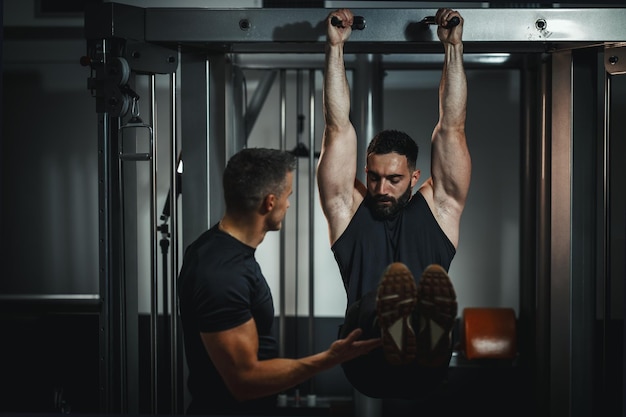 Снимок мускулистого парня в спортивной одежде, тренирующегося с личным тренером в спортзале. Он делает приседания во время силовой тренировки.