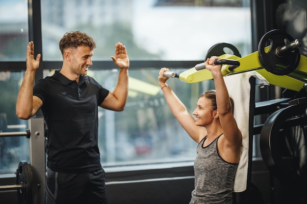 Shot of a muscular young woman in sportswear working out with personal trainer at the gym machine. She is pumping up her shoulder muscule with heavy weight.