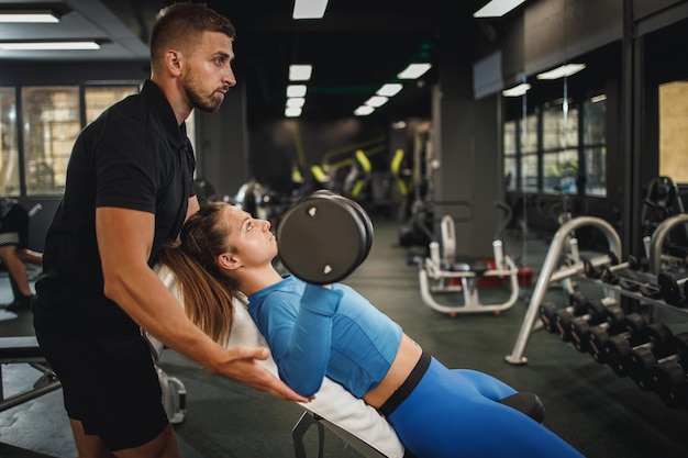 Shot of a muscular young woman in sportswear working out with personal trainer at the gym. She is pumping up her muscle with dumbbell.