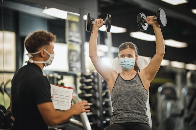 Covid-19 전염병 동안 체육관에서 개인 트레이너와 함께 운동하는 보호 마스크를 쓴 근육질의 젊은 여성의 사진. 그녀는 아령으로 근육을 펌핑하고 있습니다.