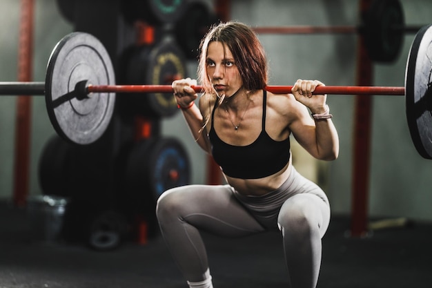 체육관에서 힘든 훈련을 하는 근육질의 젊은 여성의 샷. 그녀는 무거운 무게로 스쿼트 운동을 하고 있습니다.