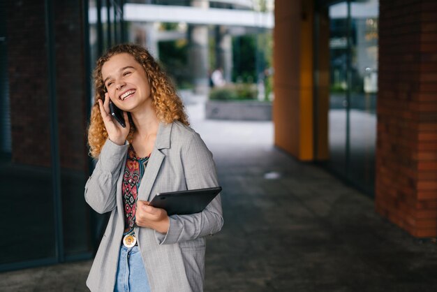 写真 建物の外に立っているラップトップ コンピューターを運ぶ彼女の携帯電話で誰かと話している笑顔の若い女性のショット コピー スペース 屋外で勉強している学生 テキスト用のスペース