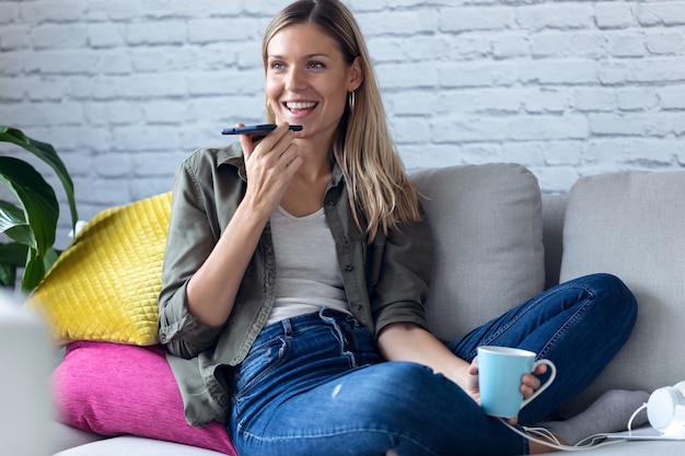 Фото Выстрел из красивой молодой женщины разговаривает по мобильному телефону с руками, выпивая чашку кофе на диване у себя дома.