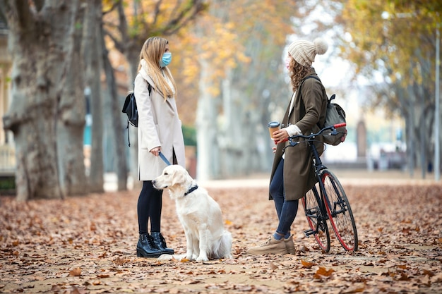 Фото Выстрел из двух красивых друзей в хирургической маске, говорящих во время прогулки со своей собакой и велосипедом в парке осенью. концепция социальной дистанции.