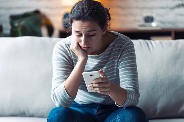 写真 自宅の居間でソファに座って携帯電話を使って心配している若い女性のショット。