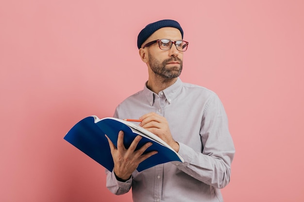 Shot of pensive unshaven man concentrated aside thinks about what to write Handsome male architect holds book and crayon underlines information for project isolated over pink background