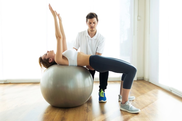 Shot of physiotherapist helping patient to do exercise on fitness ball in physio room.