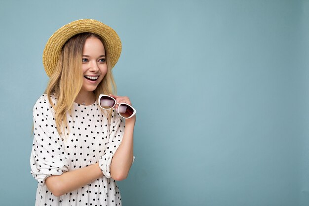 Colpo di una giovane donna bionda piuttosto positiva che indossa un cappello di paglia vestito estivo e occhiali da sole eleganti isolati su una parete di fondo blu che guarda di lato