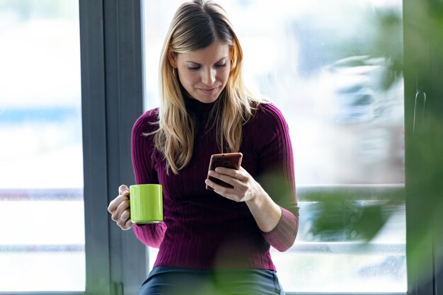 Shot of pretty young business woman sending messages with mobile phone while drinking a cup of coffee in the office.