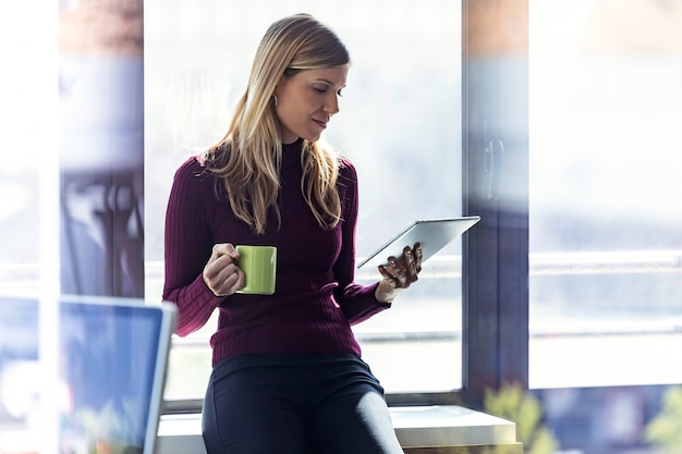 Shot of pretty young business woman using her digital tablet and holding a cup of coffee while sitting next to the window in the office.