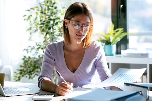 Shot of pretty young business woman writing some notes while consulting some documents in the office at home.