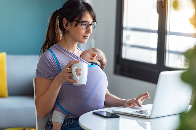 Colpo di bella giovane madre con il suo bambino in fascia che beve caffè mentre si lavora con il computer portatile a casa.