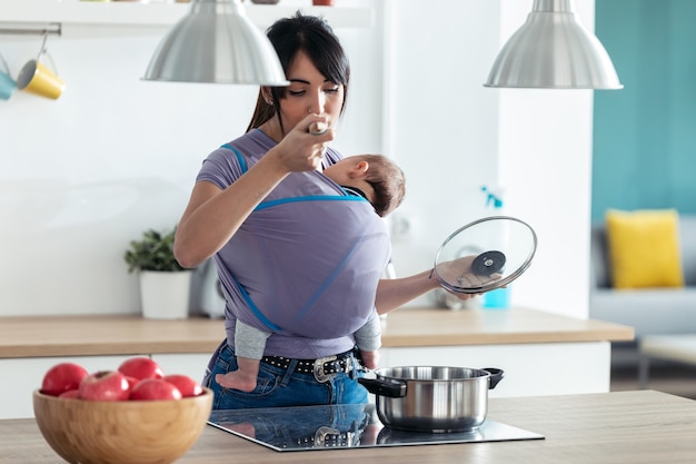 自宅のキッチンで料理をしながら、スリングで食べ物を味わう小さな赤ちゃんを持つかなり若い母親のショット。