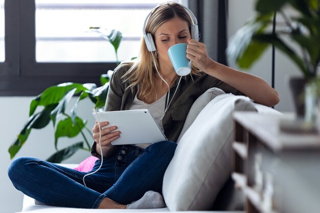 Colpo di bella giovane donna che beve una tazza di caffè mentre ascolta la musica con il suo tablet digitale sul divano di casa.