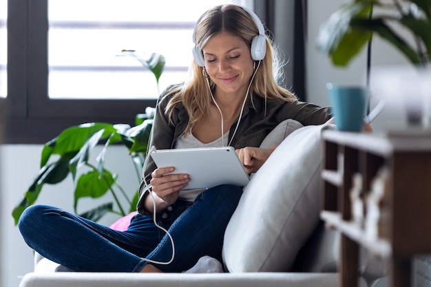 Shot of pretty young woman listening to music with headphones and her digital tablet while sitting on sofa at home.