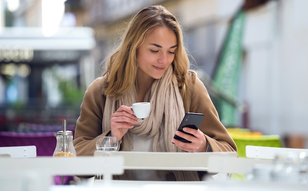 커피숍 테라스에서 커피 한 잔을 마시며 휴대폰을 사용하는 예쁜 젊은 여성의 사진.