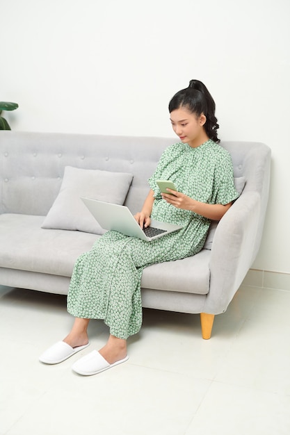 Shot of pretty young woman using her mobile phone while working with laptop sitting on sofa at home.