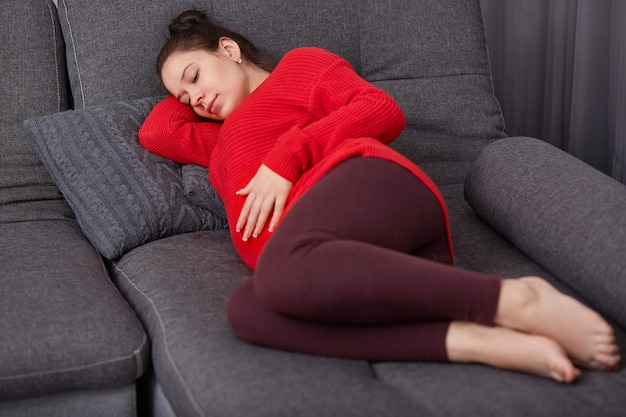 Photo shot of restful pregnant woman keeps hand on tummy, wears casual clothes, lies on comfortable sofa, going to take nap