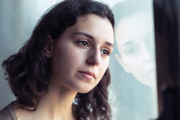 Photo shot of sad young woman crying while looking through the window at home.