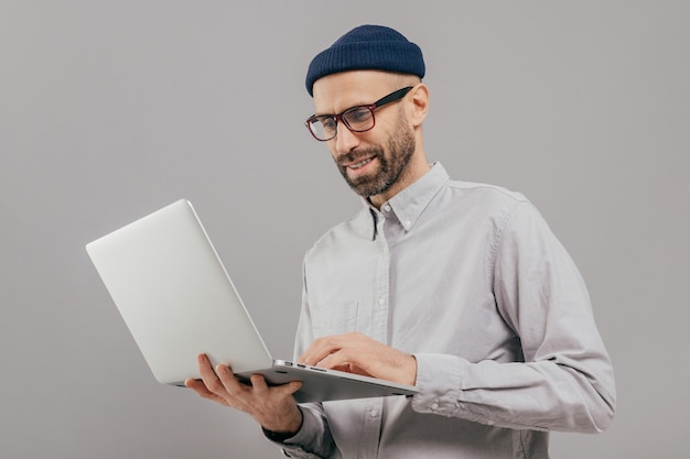 Shot of satisfied male with thick beard and mustache holds laptop computer transfers money uses online banking service sends files connected to wireless internet models over grey background