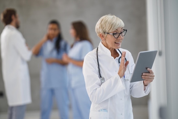 Shot of a senior female doctor making video call on a digital tablet in a hospital hallway during the Covid-19 pandemic.