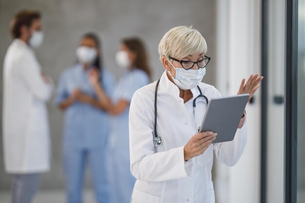 Shot of a senior female doctor with protective mask making video call on a digital tablet in a hospital hallway during the Covid-19 pandemic.