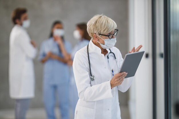 Shot of a senior female doctor with protective mask making video call on a digital tablet in a hospital hallway during the Covid-19 pandemic.