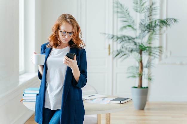 Shot of serious female entrepreneur focused in mobile phone\
makes payment via banking application drinks aromatic coffee\
recieves notification wears in elegant formal suit poses\
indoor